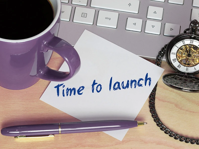 Postit mit Text: "Time to Launch" liegt halb auf Tastatur und Tisch. Kaffeehäferl, Stift und Taschenuhr sind herum drapiert.