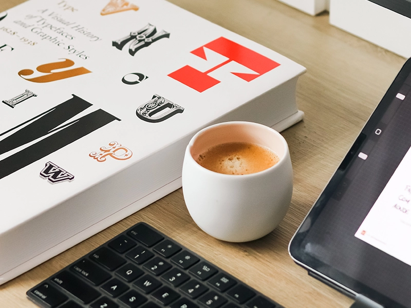 Arbeitsplatz mit Kaffeebecher in der Mitte. Danebenliegend ansatzweise zu erkennen: Tastatur, aufgestelltes Tablet, Buch zum Thema Typografie